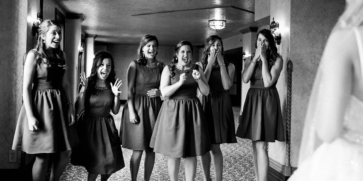 Bridesmaids in short dark dresses laughing and gasping in a hallway, black and white wedding photo